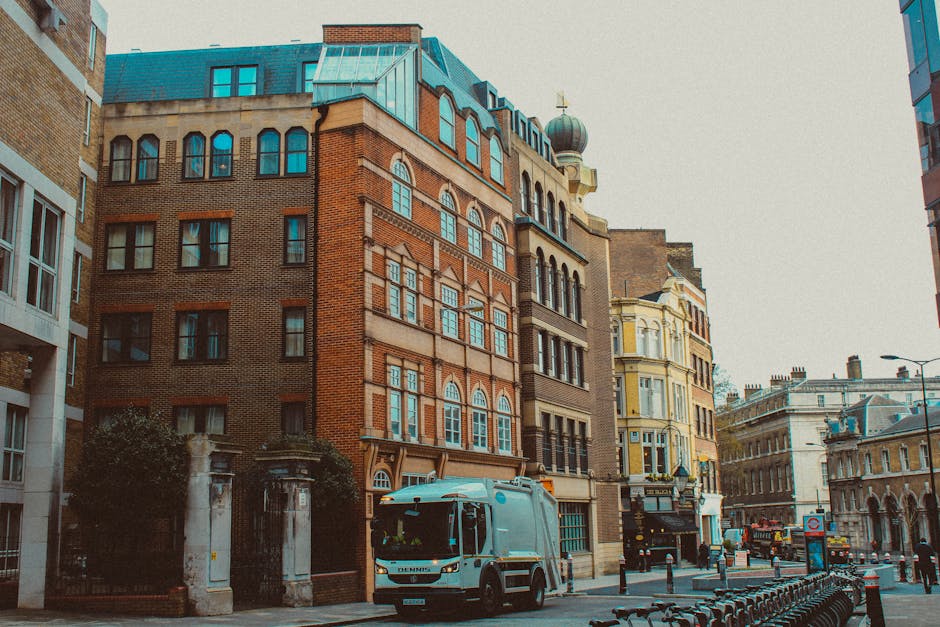 A city street scene featuring a collection of historic and modern multi-storey buildings with varied architectural styles and materials, including red brick and stucco finishes. The prominent building in the centre has large, arched windows with white frames, decorative brickwork, and a rounded, turret-like roof detail topped with a dome and a weather vane. Adjacent structures include a yellow-painted building with detailed window frames and a nearby beige and white facade. In the foreground, a white waste collection vehicle with a streamlined design is parked on the street, situated near a row of black metal bicycle racks and pedestrian bollards. The street scene is illuminated by natural daylight, with a clear sky and some available ambient light, capturing a typical urban environment where waste collection services like Rubbish Removal Hammersmith might operate or provide private waste handling solutions, especially in historic or busy commercial areas. Overall, the scene reflects a typical city street with a mix of architectural styles and waste management activity, emphasizing the importance of professional rubbish removal in maintaining city cleanliness.