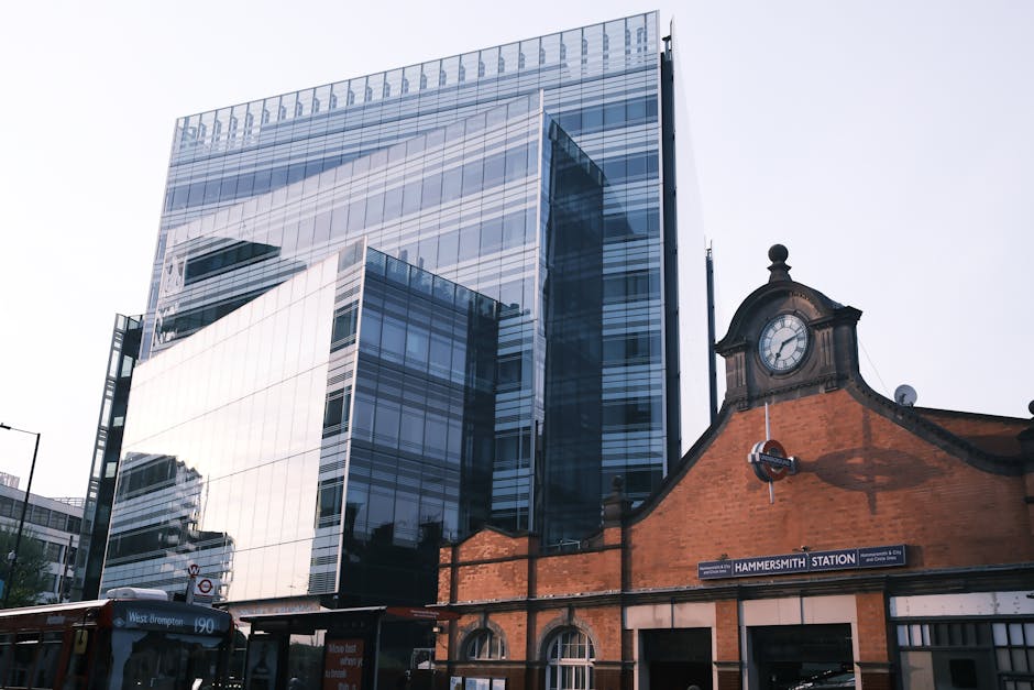 A modern multi-story office building with a glass exterior reflecting the surrounding cityscape, situated behind a historic brick station building with a rounded clock face and a blue station sign reading 'Hammersmith Station'. In the foreground, part of a bus with the route number 190 is visible at the bus stop. The scene is captured during daylight with an overcast sky, and the environment includes urban infrastructure such as street lamps and signage, creating a contrast between contemporary architecture and traditional railway station design. The image illustrates the setting around a major transportation hub, potentially related to the topic of alternative waste handling and rubbish removal options in busy urban areas.