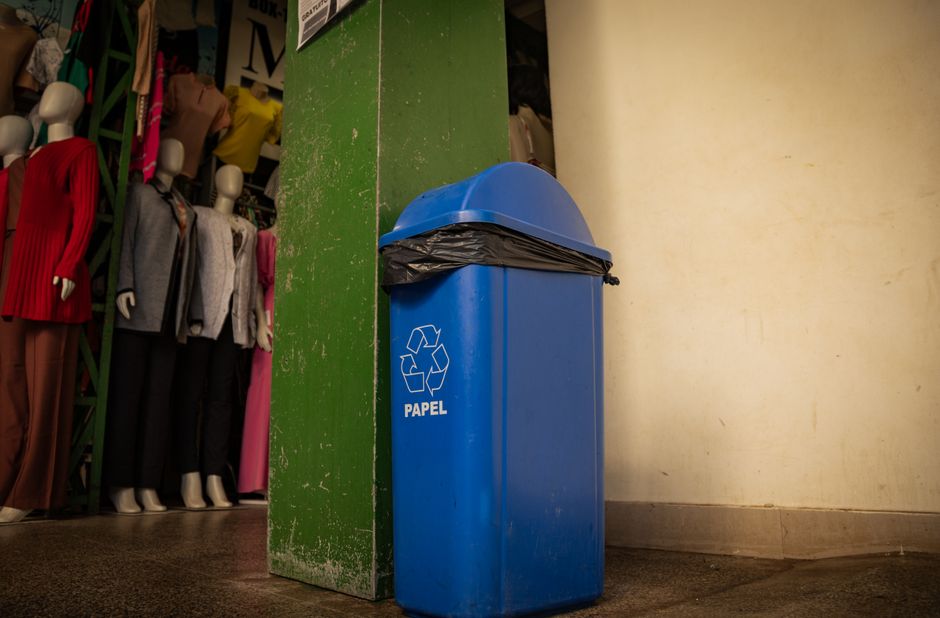 A large blue recycling bin positioned on a paved sidewalk in front of a multi-story white brick building with six evenly spaced rectangular windows that have clear glass panes reflecting the blue sky and clouds. The bin is filled beyond capacity, with several black rubbish bags stacked on top and spilling over the edges, indicating an overflowing waste collection. The bin bears the logo 'renewi' in white text. Surrounding the bin is a patch of grass and small plants directly behind it, with a neat concrete pathway at the front. The environmental context suggests an urban or suburban area where private waste disposal services, such as those provided by Rubbish Removal Hammersmith, are used to manage rubbish collection separate from municipal services. The overall scene emphasizes on-site waste storage and indicative of alternative waste handling methods often used by commercial or residential clients in Hammersmith seeking independent rubbish removal solutions.