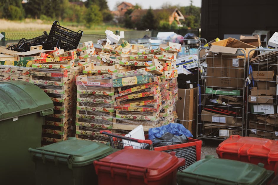 An outdoor scene showing large stacks of empty cardboard fruit and vegetable boxes arranged in rows, primarily lying flat and upright. The boxes are decorated with colourful labels and branding, featuring images and text promoting fresh produce. Surrounding the stacks are various waste containers, including green and red wheelie bins with closed lids, and metal cages filled with brown cardboard boxes, packing materials, and smaller packaging items. To the right, a metal trolley filled with more cardboard boxes and waste materials is visible. The setting appears to be a designated area for waste collection or on-site clearance, with a background of greenery and distant buildings under natural daylight. The scene emphasizes an organized but cluttered environment typical of private rubbish handling, supported by waste removal services like Rubbish Removal Hammersmith, aligning with the theme of alternative waste disposal methods outside local authority collection.