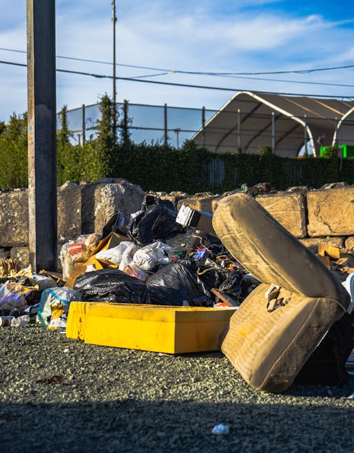A pile of mixed waste material including black plastic garbage bags, a yellow plastic storage container, a discarded beige upholstered armchair with visible fabric texture, and various miscellaneous debris scattered on a gravel surface. The waste is positioned against a low stone wall made of roughly cut, weathered stones, with some items leaning or partially overlapping each other. In the background, there is a fence with foliage behind it, and a structure with a curved roof, possibly a sports pavilion or outdoor shelter, under a bright blue sky with scattered clouds. The scene is outdoors, indicating an area where rubbish removal services from Rubbish Removal Hammersmith are often utilized for private waste collection or on-site clearance, aligned with alternative waste handling options outside local authority collection.
