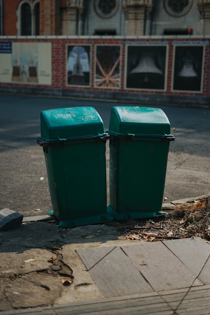 An outdoor scene showing large stacks of empty cardboard fruit and vegetable boxes arranged in rows, primarily lying flat and upright. The boxes are decorated with colourful labels and branding, featuring images and text promoting fresh produce. Surrounding the stacks are various waste containers, including green and red wheelie bins with closed lids, and metal cages filled with brown cardboard boxes, packing materials, and smaller packaging items. To the right, a metal trolley filled with more cardboard boxes and waste materials is visible. The setting appears to be a designated area for waste collection or on-site clearance, with a background of greenery and distant buildings under natural daylight. The scene emphasizes an organized but cluttered environment typical of private rubbish handling, supported by waste removal services like Rubbish Removal Hammersmith, aligning with the theme of alternative waste disposal methods outside local authority collection.