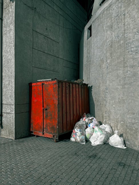 A red metal refuse container with visible rust and peeling paint is positioned against a grey concrete wall, located in a narrow urban alleyway beside a dark green building with smooth, flat cladding. The container is partially open on the top, and several clear plastic garbage bags filled with mixed waste are piled around its base, some leaning against the container and others resting directly on the textured paving surface. The bags appear loosely knotted, and waste contents, such as plastic bottles and packaging, are partially visible through the thin plastic. Behind the container, the alley extends and is partially shaded, with a small rectangular opening in the building wall providing minimal natural light. This scene illustrates an area designated for waste collection that could be associated with independent rubbish disposal services, such as those provided by private waste management companies, highlighting the importance of responsible rubbish removal and debris clearance in urban environments.