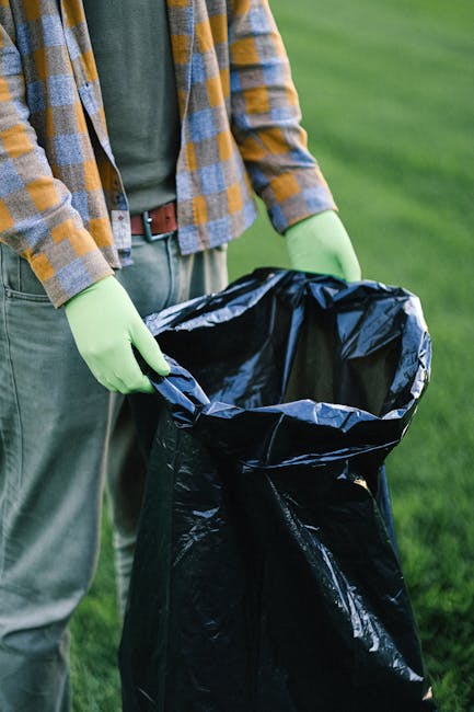 A person wearing a yellow and grey checkered shirt over a green top is holding open a black rubbish bag with bright green gloves, preparing for waste disposal. The bag is made of shiny plastic material, and the person's hands are positioned at the opening, ready to either fill or seal it. The scene is set outdoors on a green grassy area, suggesting a garden or park environment. The background features blurred greenery, indicating a natural, open space. The image emphasizes the importance of proper waste handling and the use of protective gloves, consistent with professional rubbish removal services such as those offered by Rubbish Removal Hammersmith, which facilitate alternative waste disposal options beyond local authority collections.