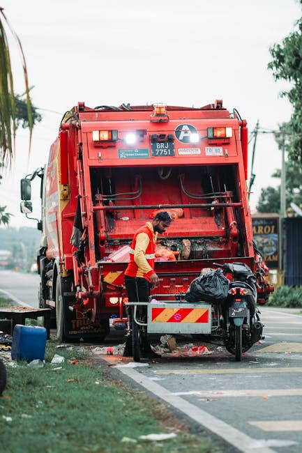 A municipal waste collection worker wearing a high-visibility yellow and orange uniform is operating the back of a large red rubbish truck on a paved street. The truck is equipped with hydraulic arms and a large, rectangular rear loading area with a black trash bag placed nearby. The worker is focused on loading waste, with debris and small trash items visible on the ground around the truck’s rear. The scene takes place during daylight under overcast skies, with trees and utility poles visible in the background. The environment appears to be an urban or suburban roadside area, with a section of curb and roadside grass in the foreground, emphasizing the context of waste collection and disposal, which relates to private rubbish removal and alternative waste handling options often provided by services like Rubbish Removal Hammersmith.