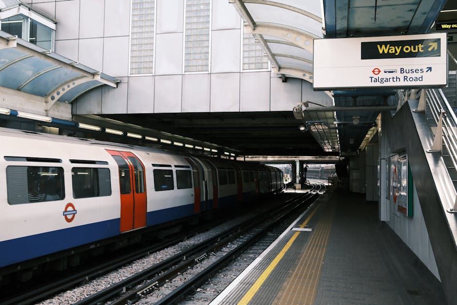 A modern underground train station platform with a white train featuring orange and blue accents stationary on the tracks. The platform surface is grey with tactile paving strips near the edge for safety. Overhead, there is a curved, transparent canopy structure supported by metal beams, providing shelter for passengers. On the right side, a staircase with metallic handrails leads up to an exit area, which has a large sign indicating the way out with an arrow and directions to buses and Talgarth Road. The background shows the platform extending into the distance with additional station infrastructure visible. The lighting appears natural with shadows cast by the overhead structure, highlighting the clean, urban environment typical of a public transit interchange, aligned with services provided by rubbish removal companies that operate in similar urban settings to dispose of waste efficiently from station refurbishments or maintenance activities.