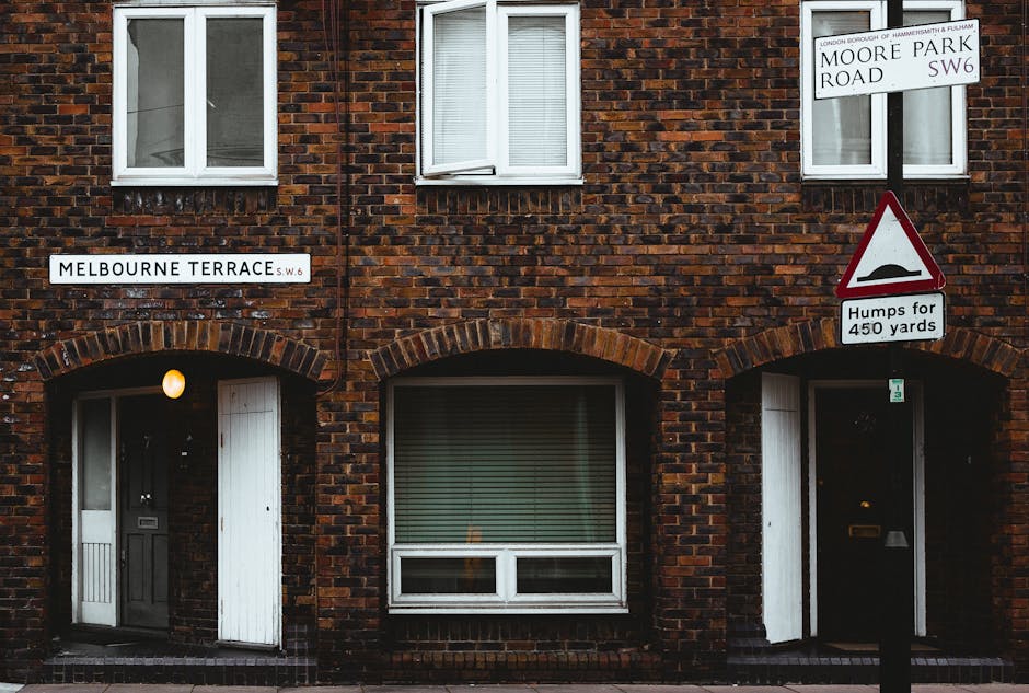 The image depicts a brick residential building on Melbourne Terrace, SW6, with two visible ground-floor entrances and three upper-floor windows. The brickwork is dark red with a slightly rough texture, and the mortar joints are prominent. Above each entrance, there are rounded brick arches, and the doorways have protective coverings, with one door painted dark and the other white, both adjacent to small light fixtures. In front of the building, there is a black metal pavement, and two street signs are mounted on wooden posts: one indicating the street name 'Melbourne Terrace,' and another showing the intersection with Moore Park Road, SW6, along with a road condition warning sign about bumps for 450 yards, featuring a black bump icon on a triangular red border. The environment is typical of an urban residential area, with minimal vegetation visible. The lighting appears to be natural daylight, creating a clear and neutral atmosphere suitable for visual inspection or documentation related to local streets and housing exterior conditions, often referenced in waste management and rubbish removal contexts such as on the website rubbishremovalhammersmith.co.uk.